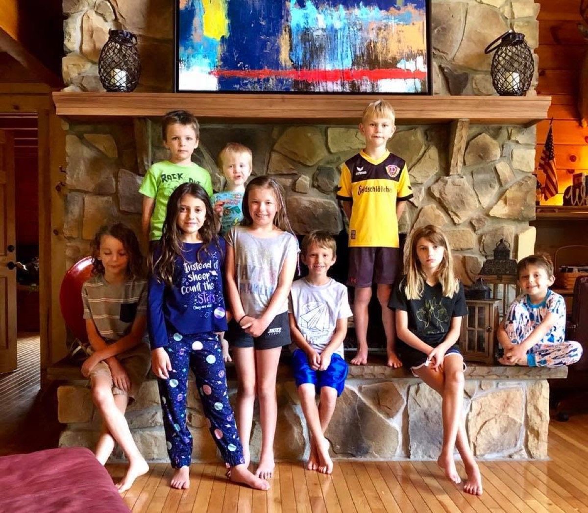 The same nine children rearranged, several perched on the stone ledges of the fireplace, the smallest sitting on the hearth at the right.