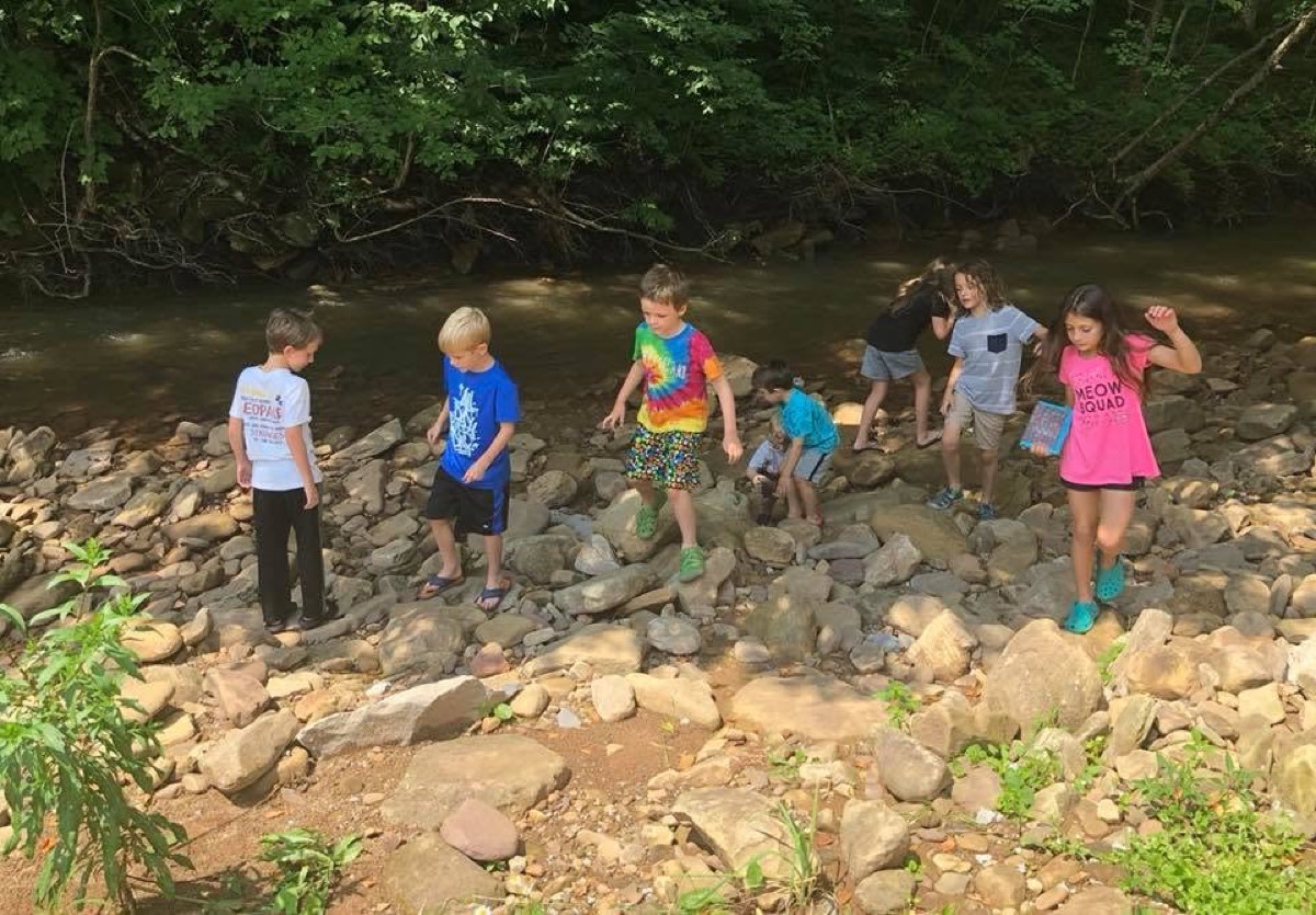 Eight children spread across a wide rocky creek bed in the woods, several crouched among the stones, one in a tie-dye shirt in the middle.