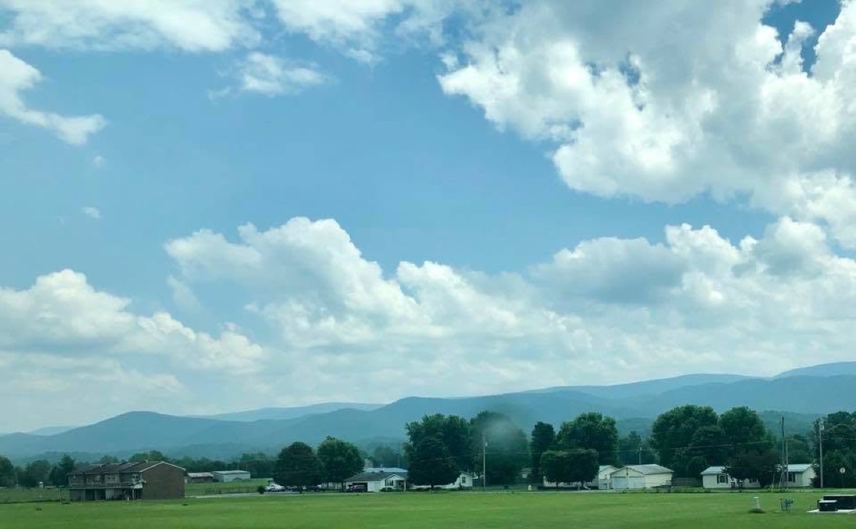 A wide green field with farm buildings along its edge under a tall bank of white cumulus cloud, a low blue ridge on the horizon.