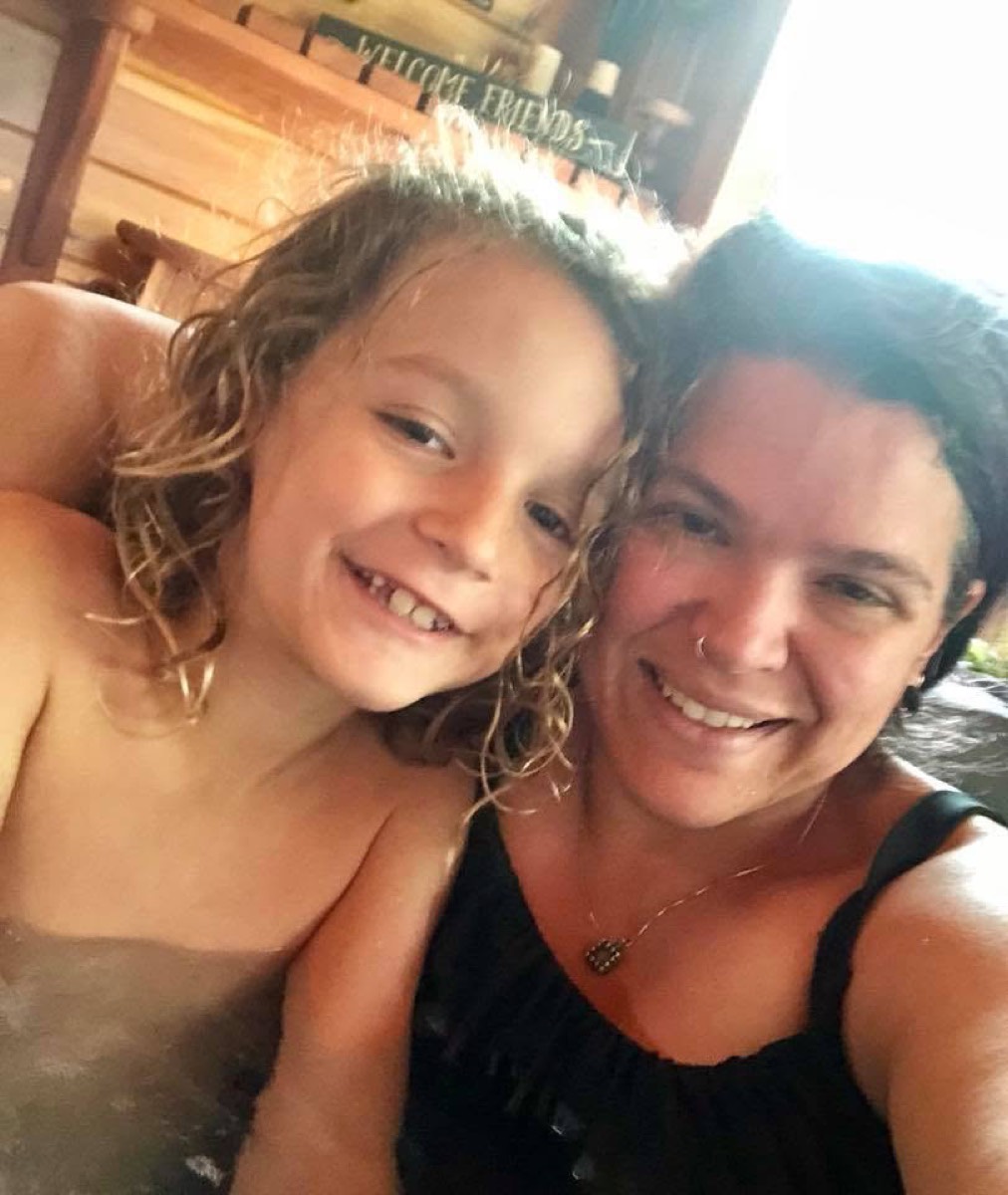 A close selfie of Jone and Mason pressed cheek to cheek, both grinning, Mason's wet hair spiked up and lit bright by the sun above them, a WELCOME FRIENDS sign on the wall behind.