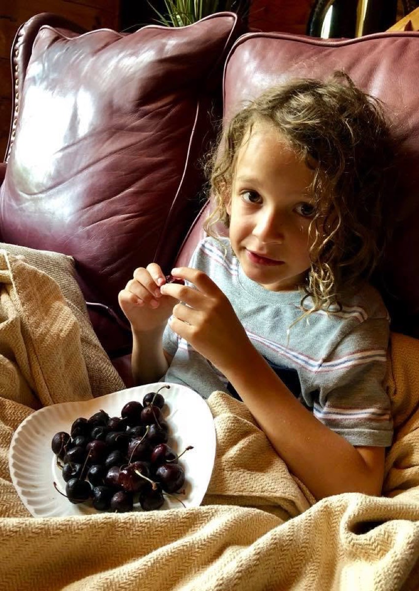 Mason sitting back on a dark red leather couch under a pale knitted blanket, holding a cherry up by the stem, a white scalloped plate piled with dark cherries beside him.