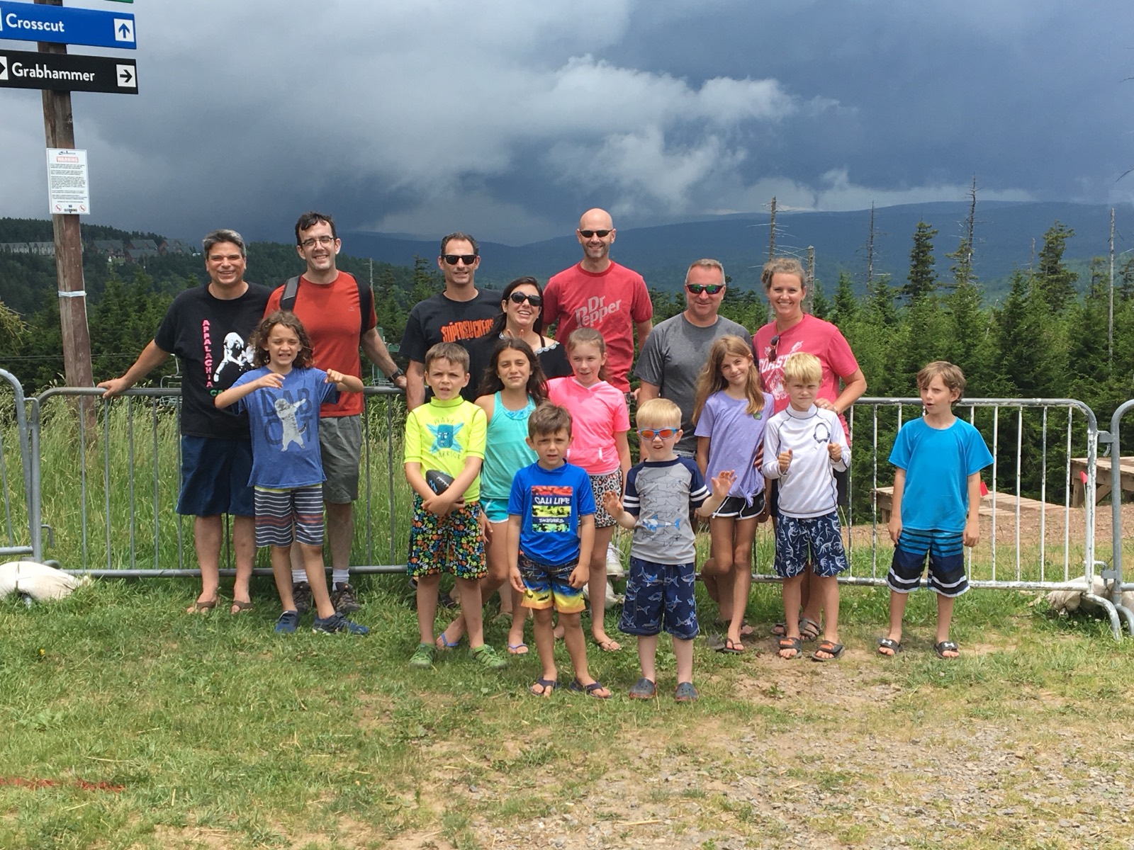 The full Chuckwagon Gang posed at a ski trail junction, adults standing in a row behind ten children, signs for the Crosscut and Grabhammer trails on a post at left and dark storm clouds massing over green mountains behind.