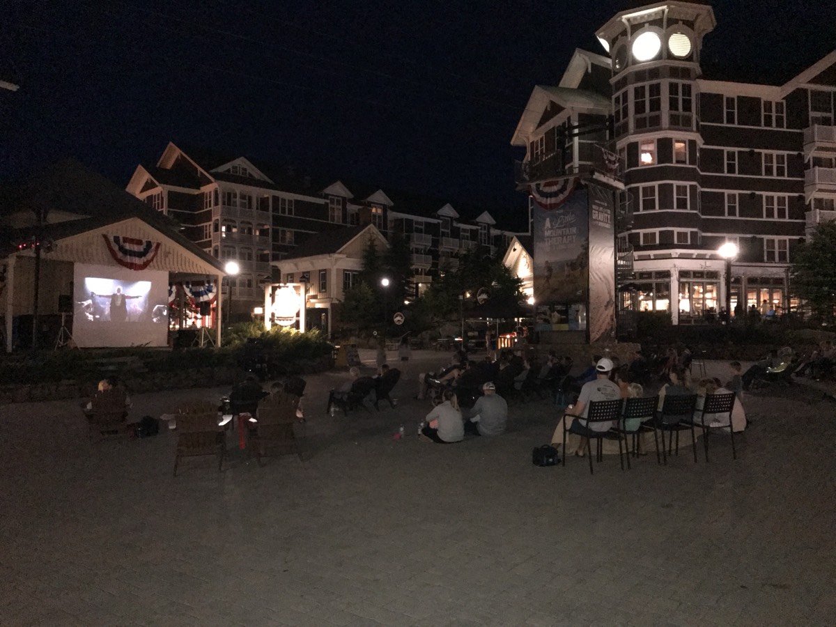 An outdoor movie screening at night in the Snowshoe village square, a lit projection screen at left, people watching from folding chairs and the pavement, illuminated lodge buildings and a clock tower behind.