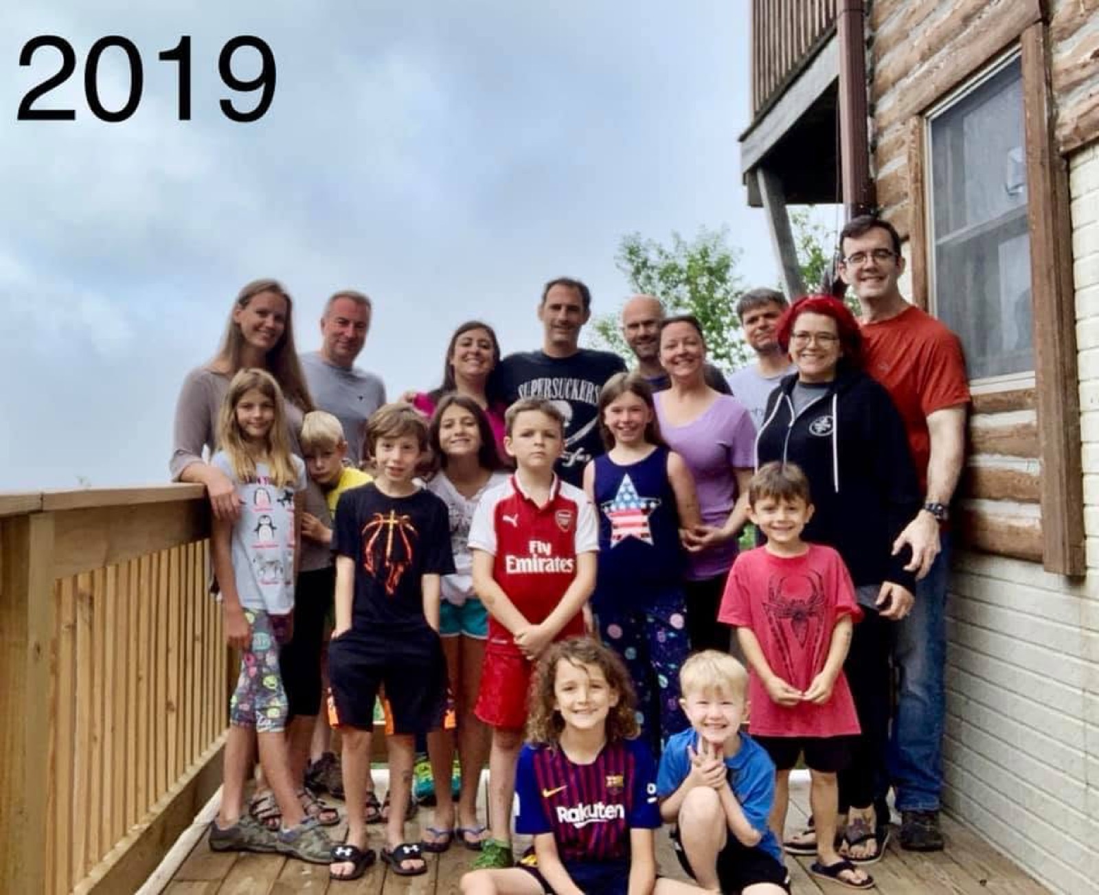 The full Chuckwagon Gang crowded onto a wooden cabin deck, nine adults standing behind nine children, log siding and a window at right, overcast sky behind; the year 2019 is written across the top left of the photo.