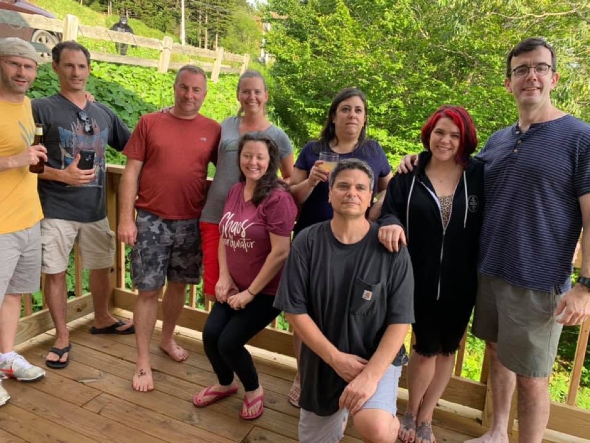 Nine adults posed together on a wooden cabin deck in the evening, some holding drinks, one man kneeling at the front, a green hillside and split-rail fence behind them.