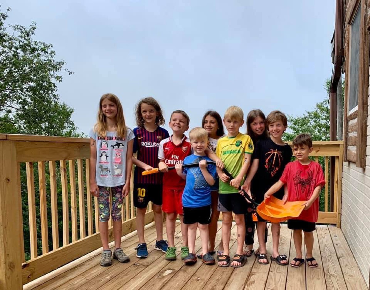 Nine children lined up along a wooden cabin deck, several holding toy blasters and one holding an orange flag, trees and overcast sky behind the railing.
