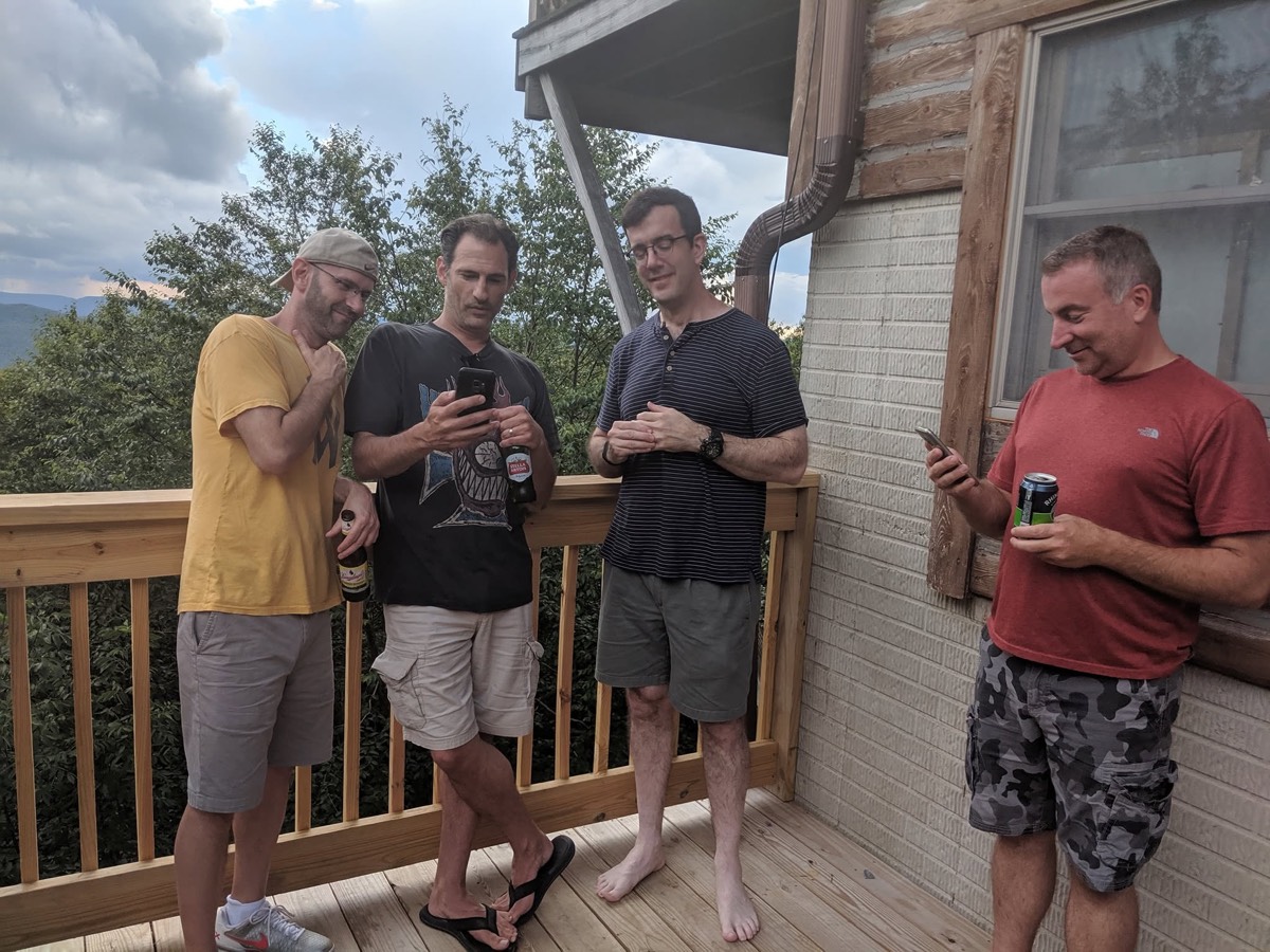 Four men standing and talking on a cabin deck holding drinks, log cabin wall and window behind them, mountains and cloud in the distance.