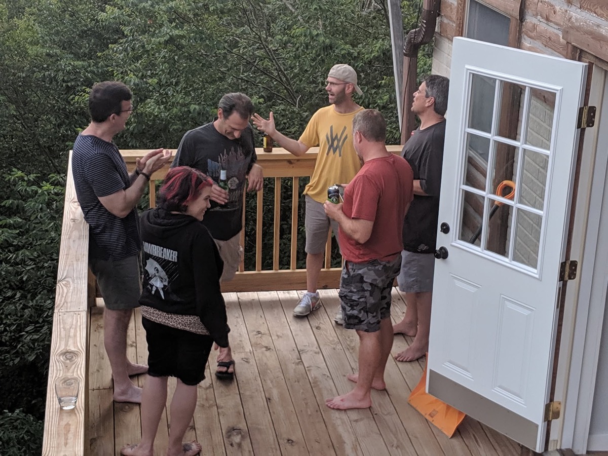 Adults gathered on a cabin deck beside an open white door, one woman with dark red hair facing away from the camera, dense green woods beyond the railing.
