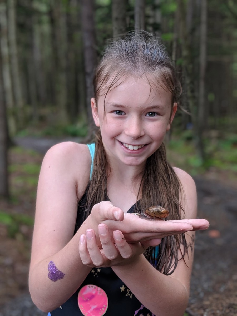 A girl with wet hair and a swimsuit smiling at the camera, a small brown snail crawling along her outstretched palm, forest path behind her.