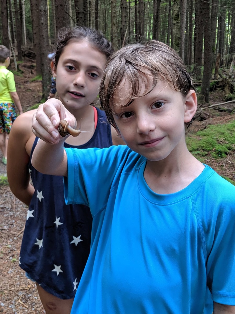 A girl in a navy star-print tank top holding a snail out toward the camera beside a boy in a bright blue shirt, both standing in the woods.
