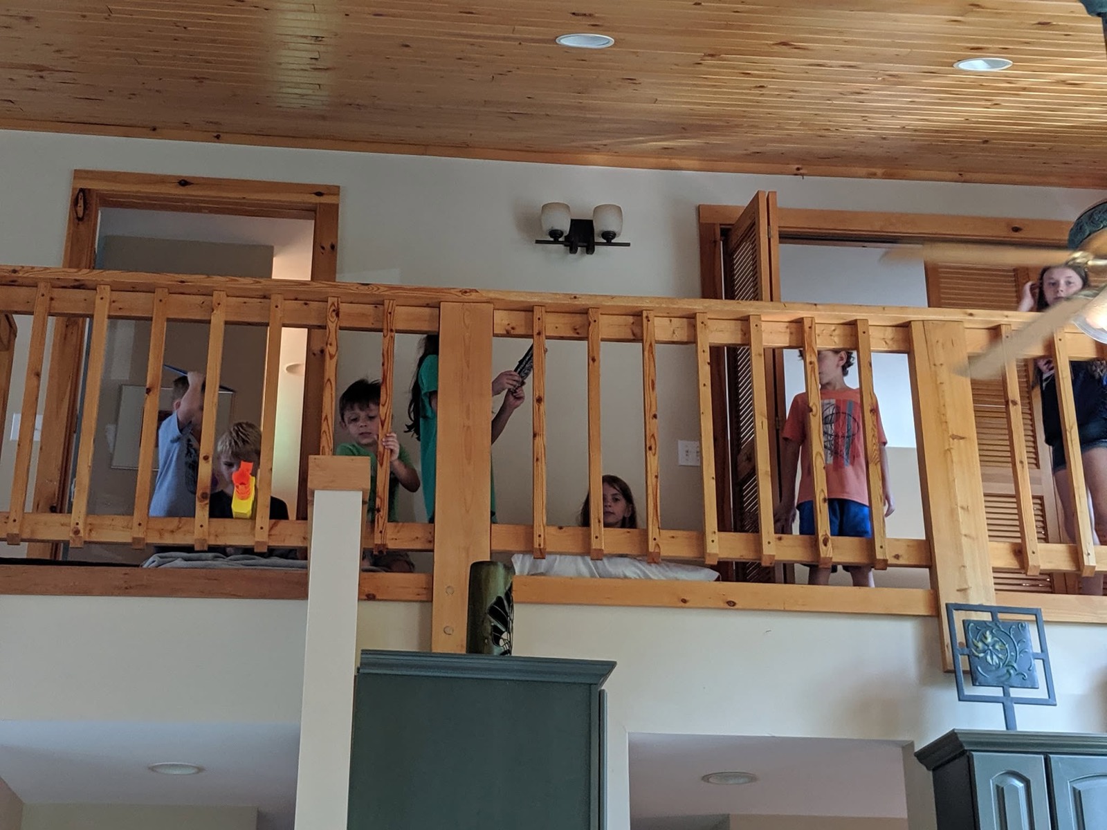 Looking up at the open second-floor landing of a log cabin, pine plank ceiling and heavy wooden railings, several children visible along the upper level.