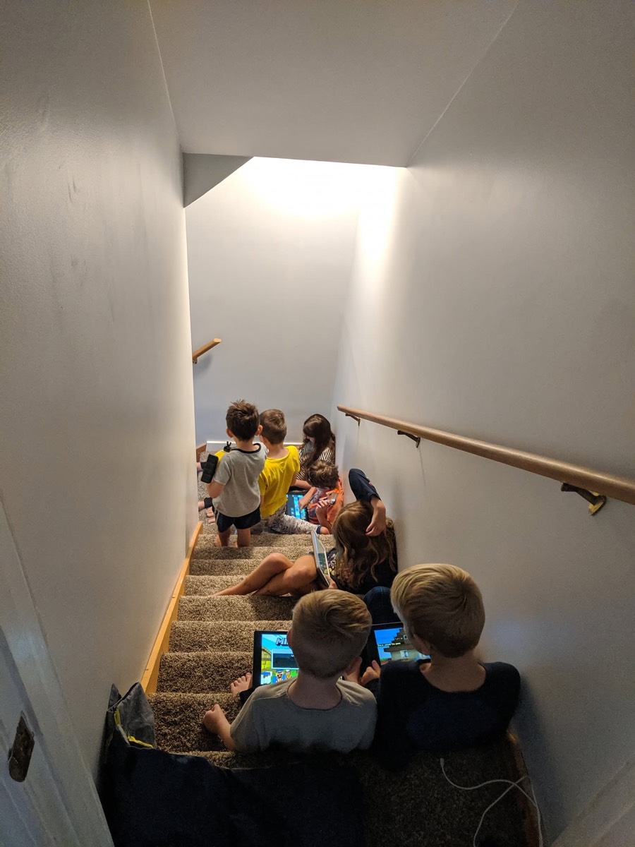 Children sitting at intervals down a carpeted staircase in the cabin, several holding tablets, daylight coming from a window above.