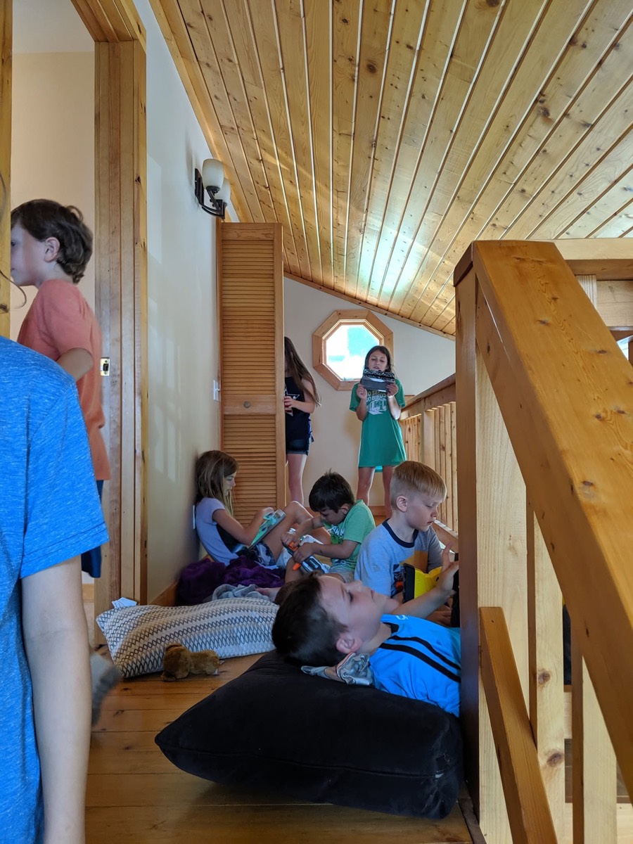 The upstairs landing of the cabin with a sloped pine ceiling and octagonal window, several children sitting and lying on cushions and pillows on the wooden floor with tablets.