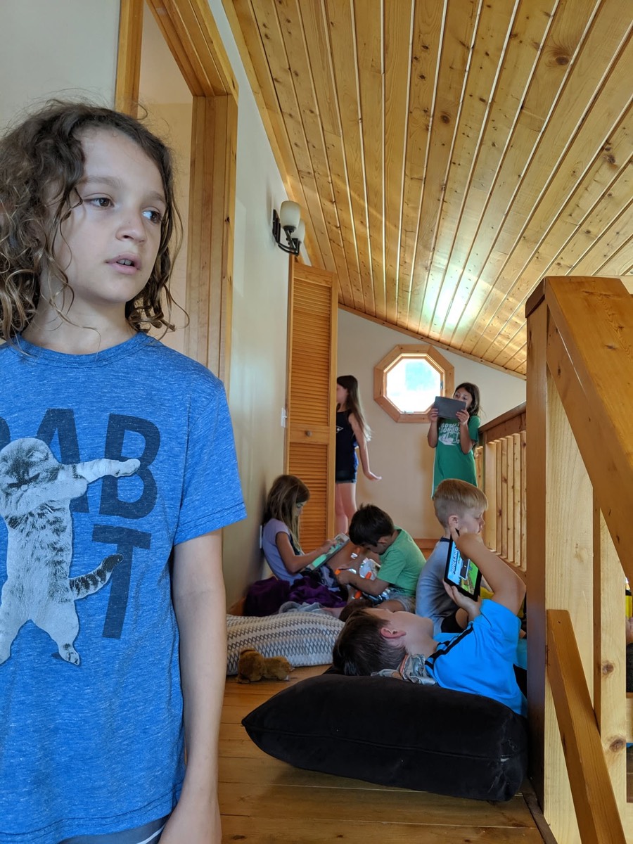 A child in a blue cat-print t-shirt standing in the foreground of the cabin's upstairs landing looking off to the side, other children on the floor behind with tablets.