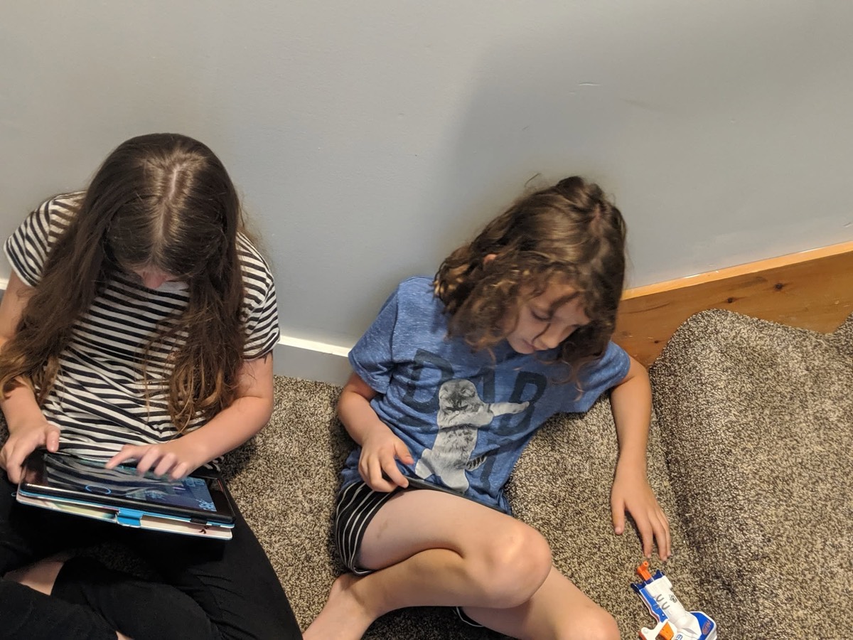 Two children slumped together asleep or nearly asleep on a couch, one still holding a tablet, a snack packet beside them.