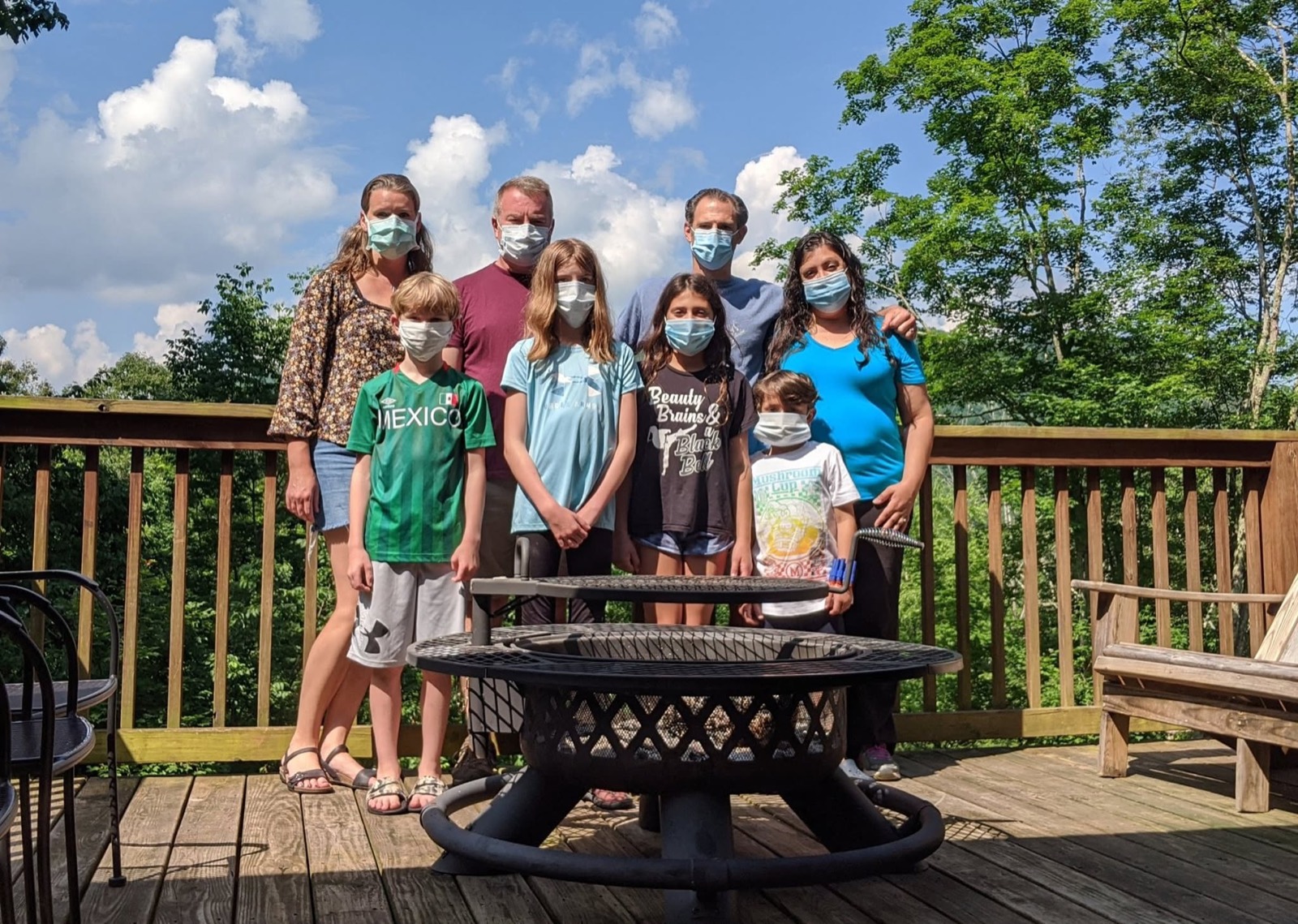 Eight people in surgical masks posed on a wooden cabin deck behind a large round fire pit, four adults standing behind four children, green trees and tall summer clouds behind them.