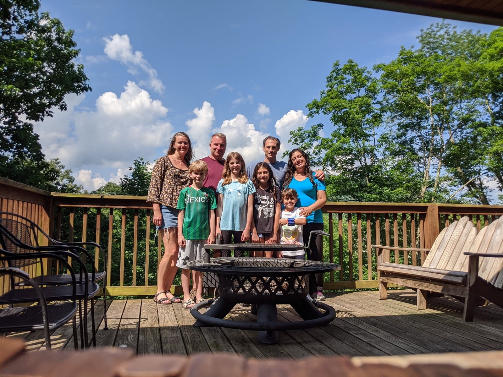 The same eight people on the same cabin deck behind the fire pit with their masks removed, all smiling at the camera under a bright blue sky with cumulus clouds.