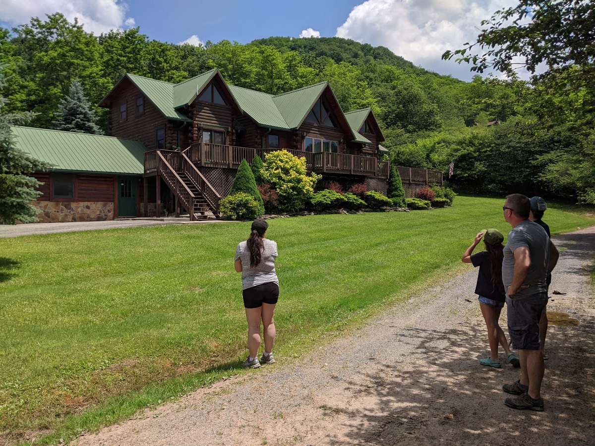 A large log cabin with a green metal roof and several gable ends, seen across a mown lawn, with four people walking up the gravel drive toward it and wooded hills behind.