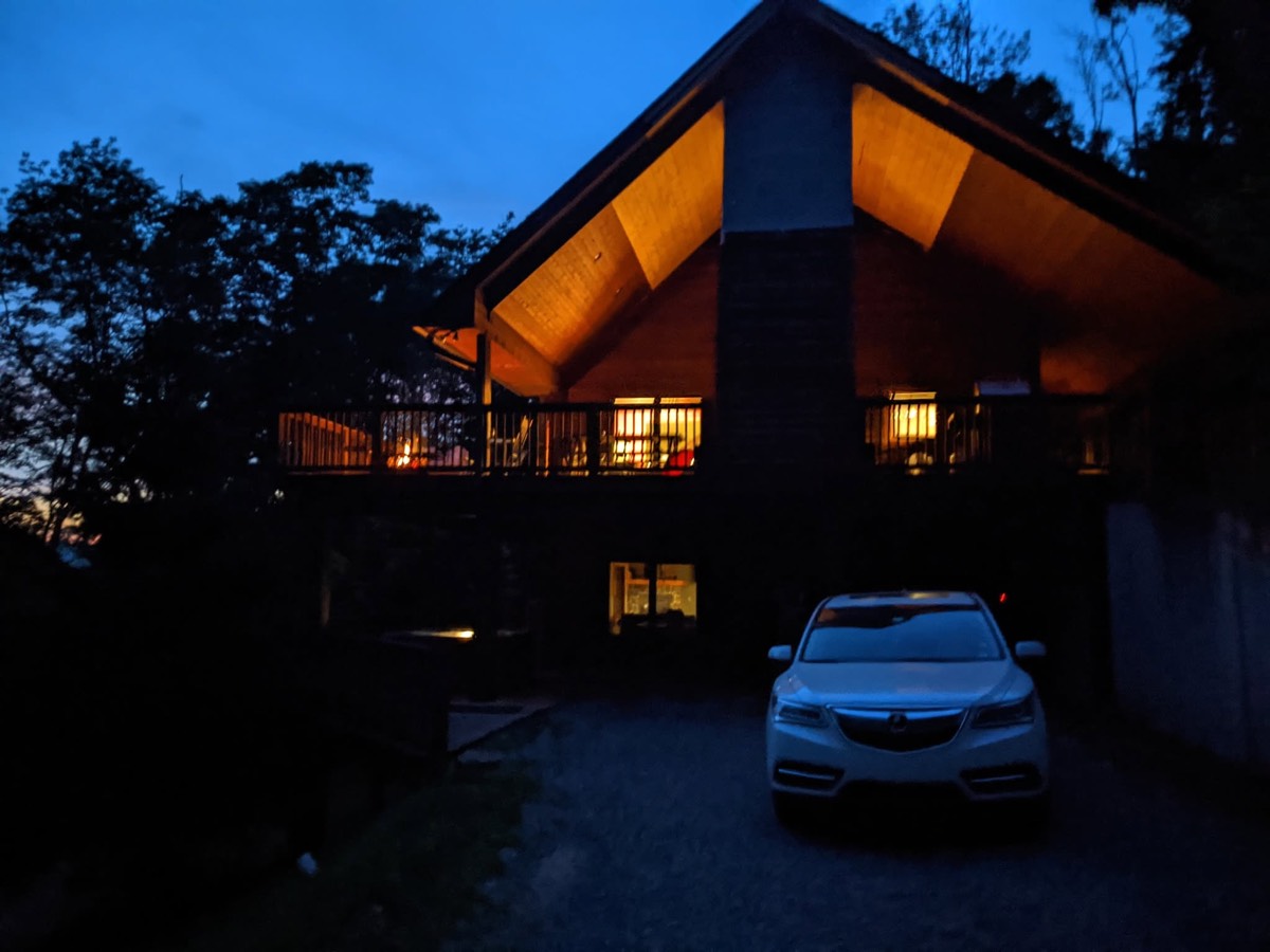 The cabin at dusk, its tall gable end and deck glowing warm yellow from inside, a car parked in the dark drive in front and deep blue sky above.