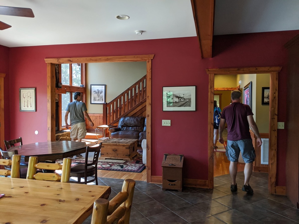 The cabin's open interior with deep red walls, a long pine dining table and chairs in the foreground, a staircase and sitting area beyond, and two people walking through the far doorways.
