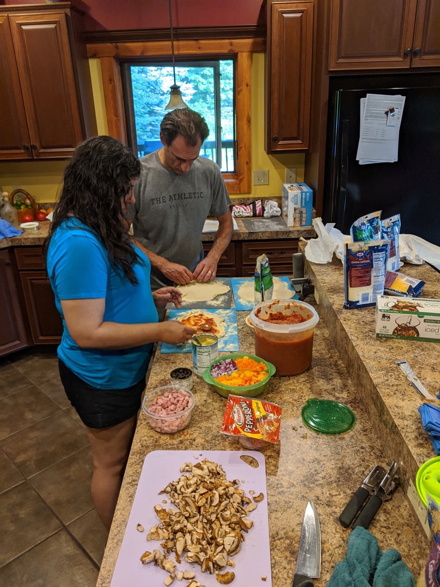 Two adults working at a granite kitchen island covered with cutting boards, chopped vegetables, snacks and bowls in a wood-cabinet cabin kitchen.