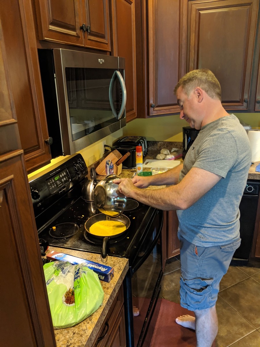 A man at the stove in the cabin kitchen tipping a pan of scrambled eggs, prep bowls and a cutting board on the counter beside him.