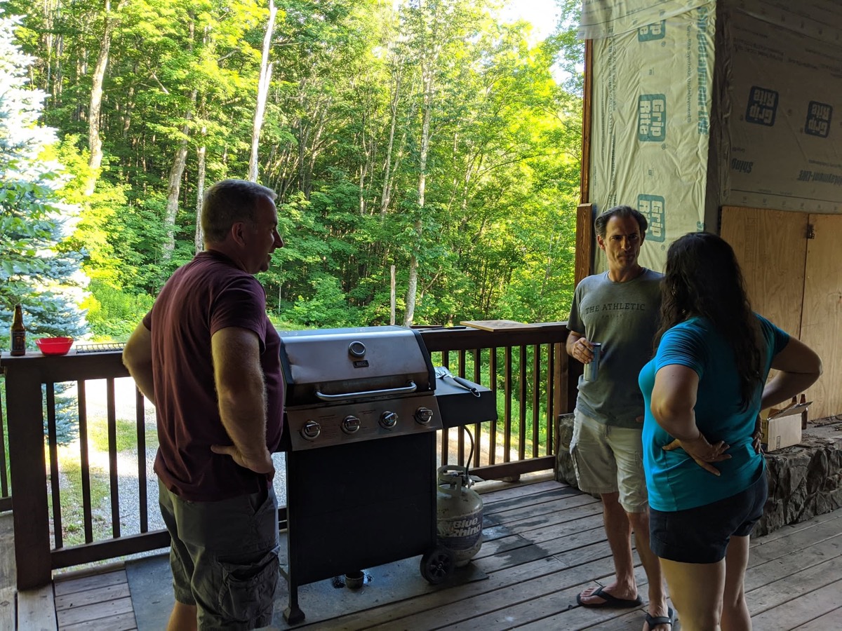 Three adults standing around a gas grill on a cabin deck, one tending it, dense green treetops and a steep wooded slope falling away behind the railing.