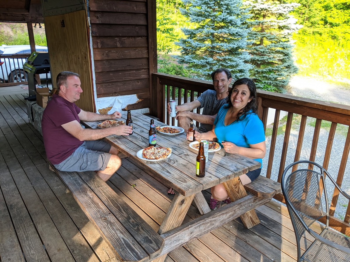 Three adults sitting at a weathered wooden picnic table on a covered porch with pizza on plates and bottles in front of them, evergreens in the sun beyond.
