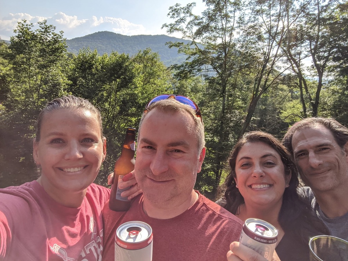 A close selfie of four adults smiling on a deck holding cans and bottles, treetops and a distant blue mountain ridge behind them.