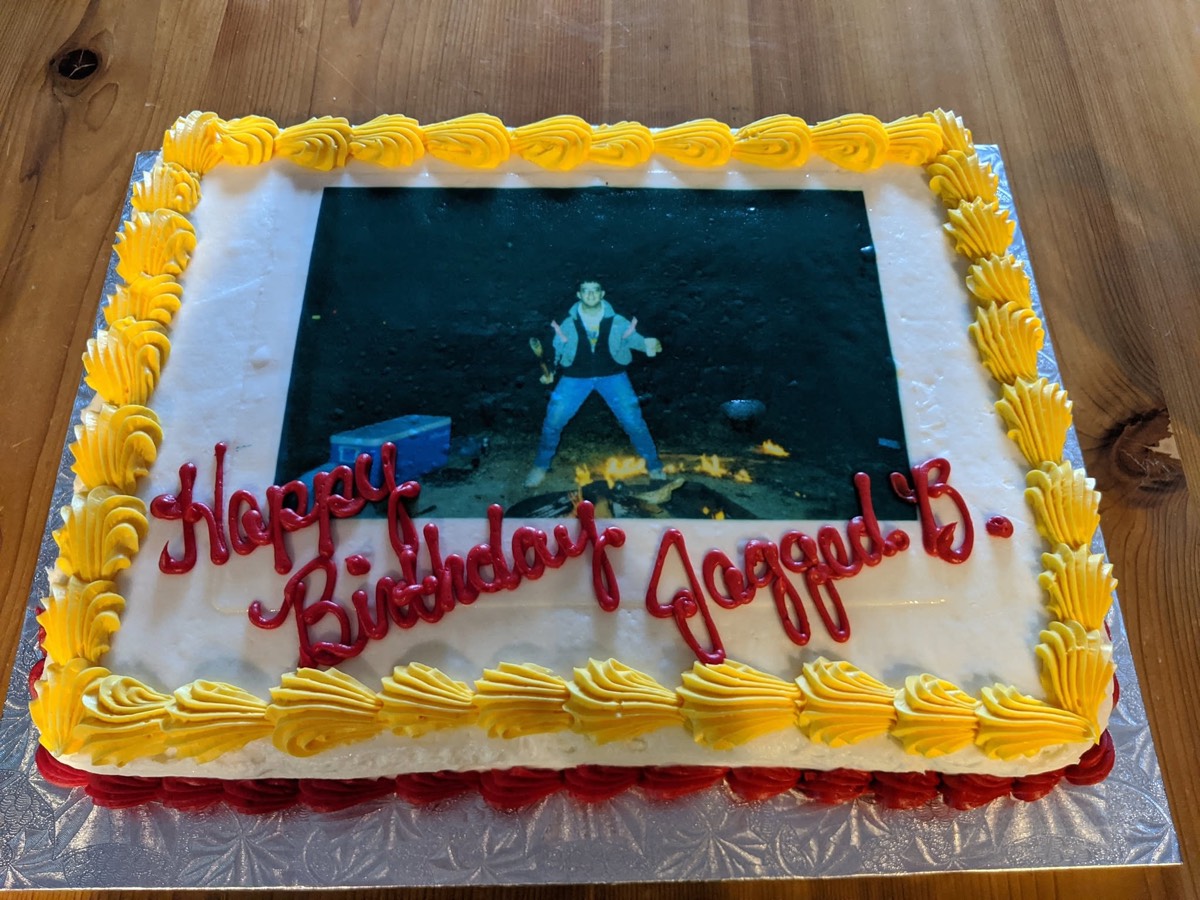 A large sheet cake with yellow icing shells around the border and Happy Birthday Jagged B piped in red, with an edible photo panel in the center showing a young man in jeans and a jacket standing arms-out over a campfire at night, a blue cooler on the ground beside him.