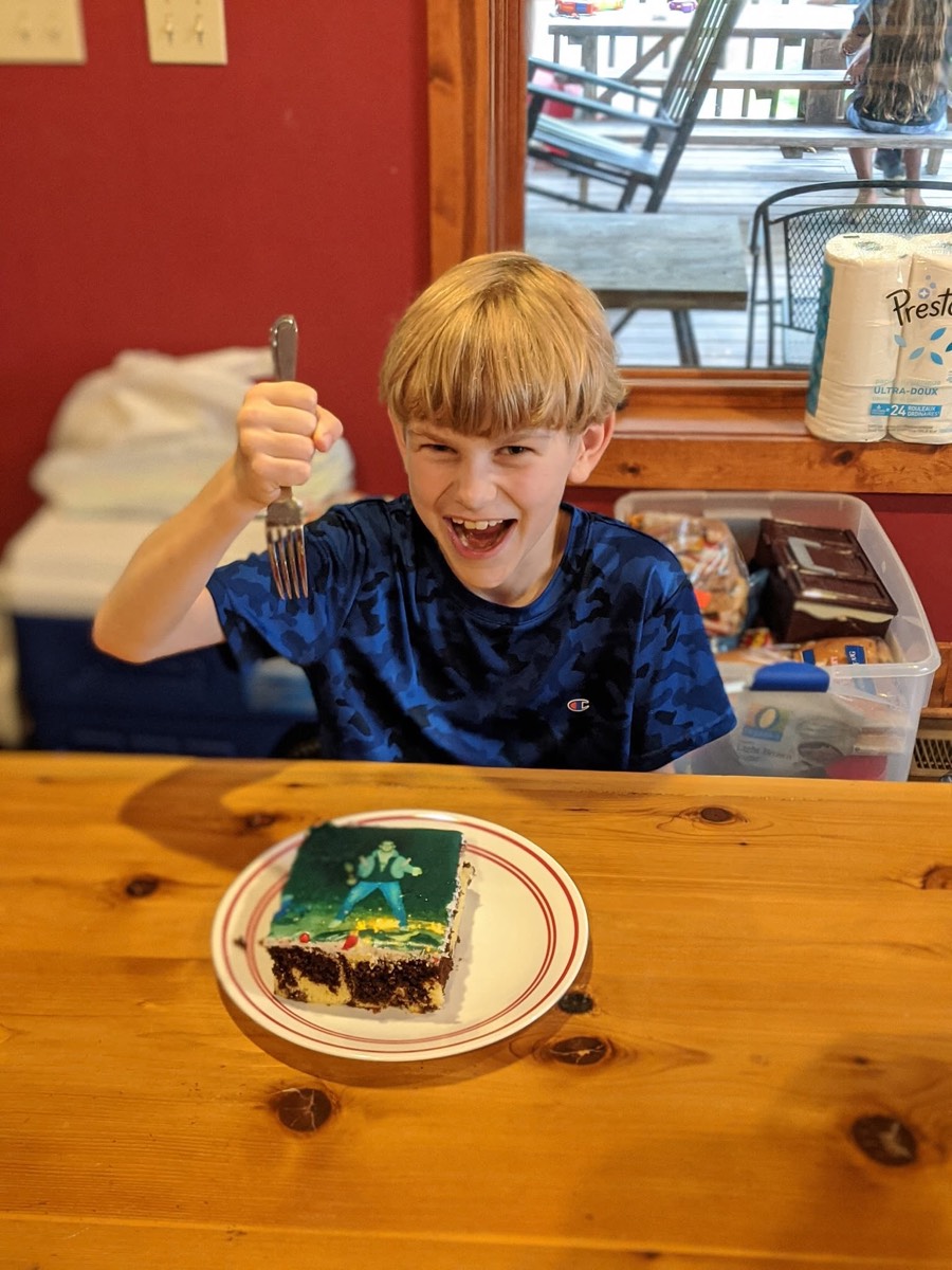 A grinning boy in a blue shirt at the cabin dining table raising a fork over a slice of the birthday cake on a plate in front of him.