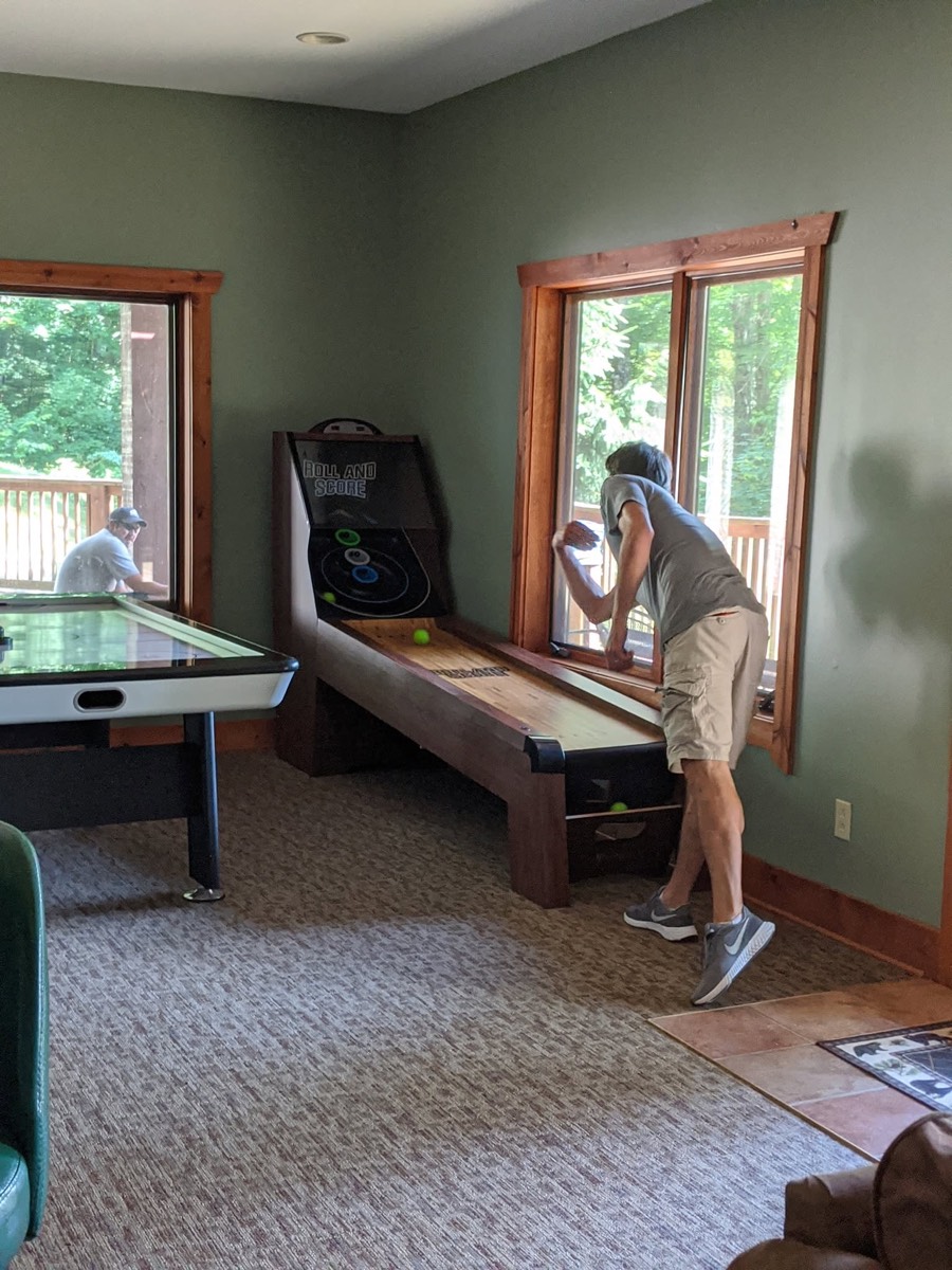 A cabin games room with an air hockey table in the foreground and a long shuffleboard table against the far wall, a man bending over the shuffleboard beside a bright window.