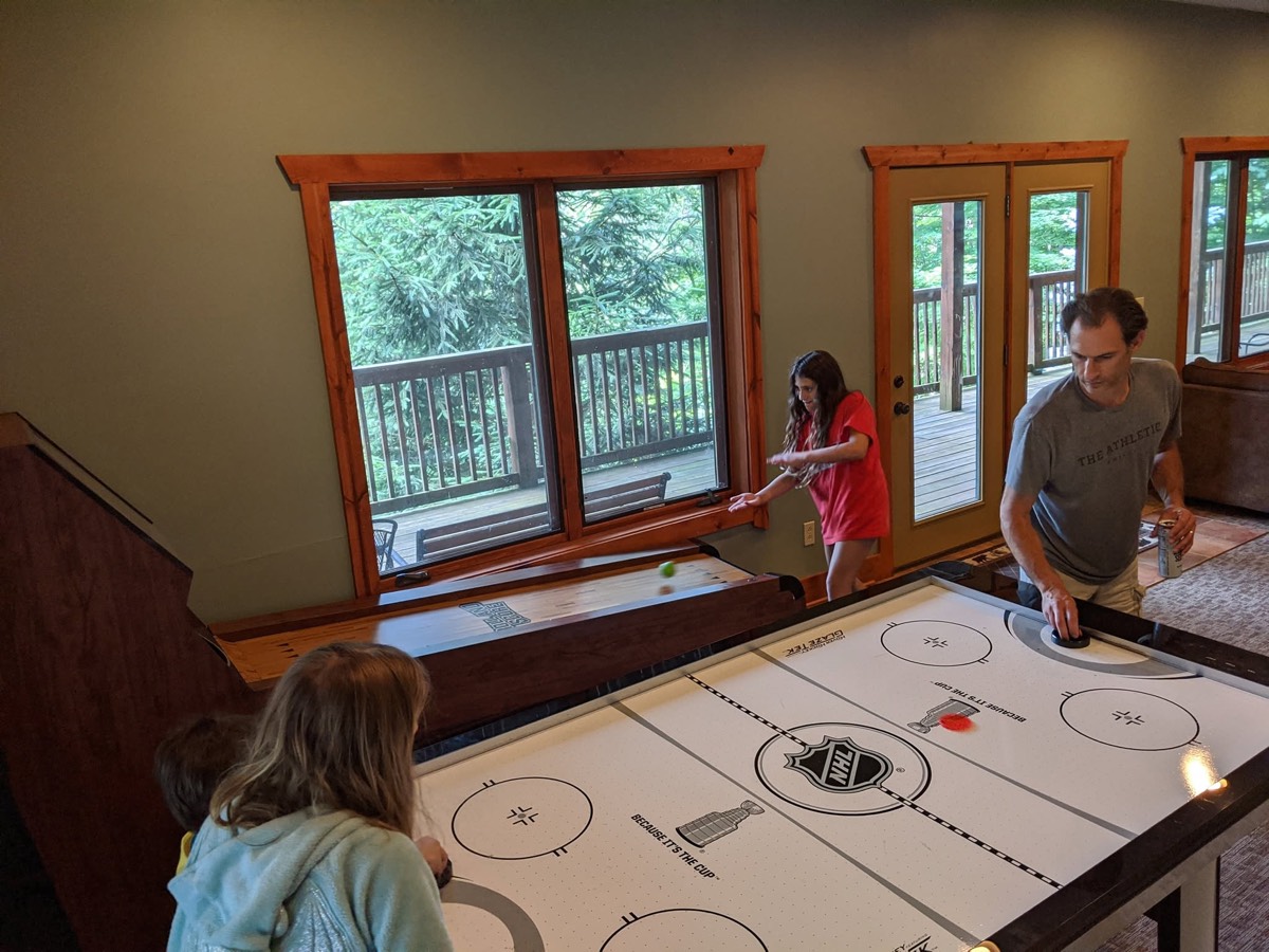 Two children and an adult playing air hockey on a table in a bright cabin room with windows and a glass door behind them.