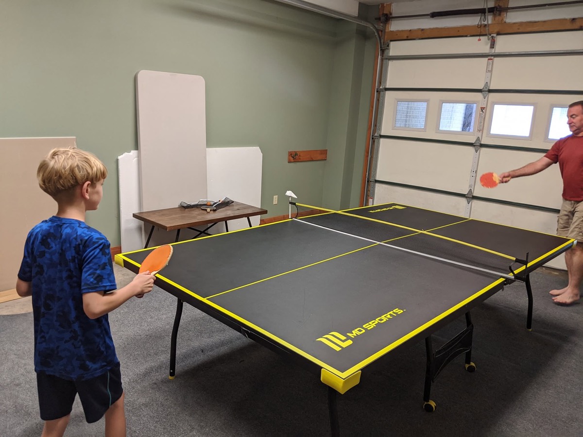 A boy in a blue shirt holding a paddle at one end of a yellow and black ping pong table set up inside a garage, an adult returning at the far end.