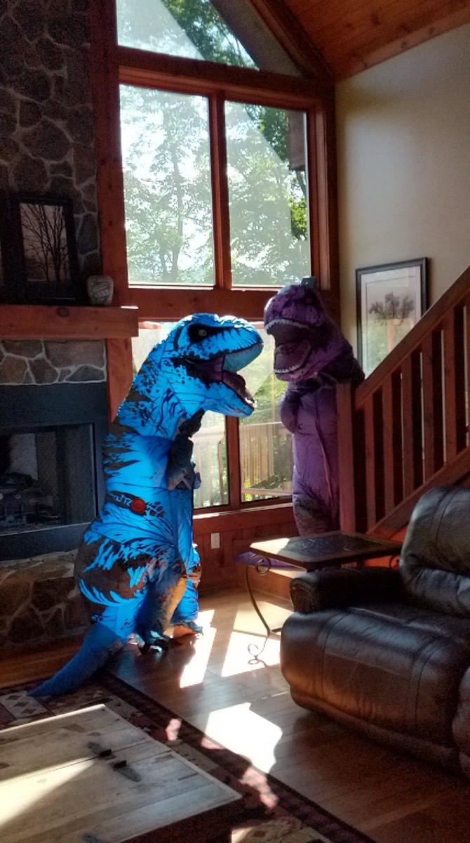 A person in a blue inflatable tyrannosaurus costume standing on a cabin floor beside the stone fireplace, with a second person in a purple costume on the staircase behind.