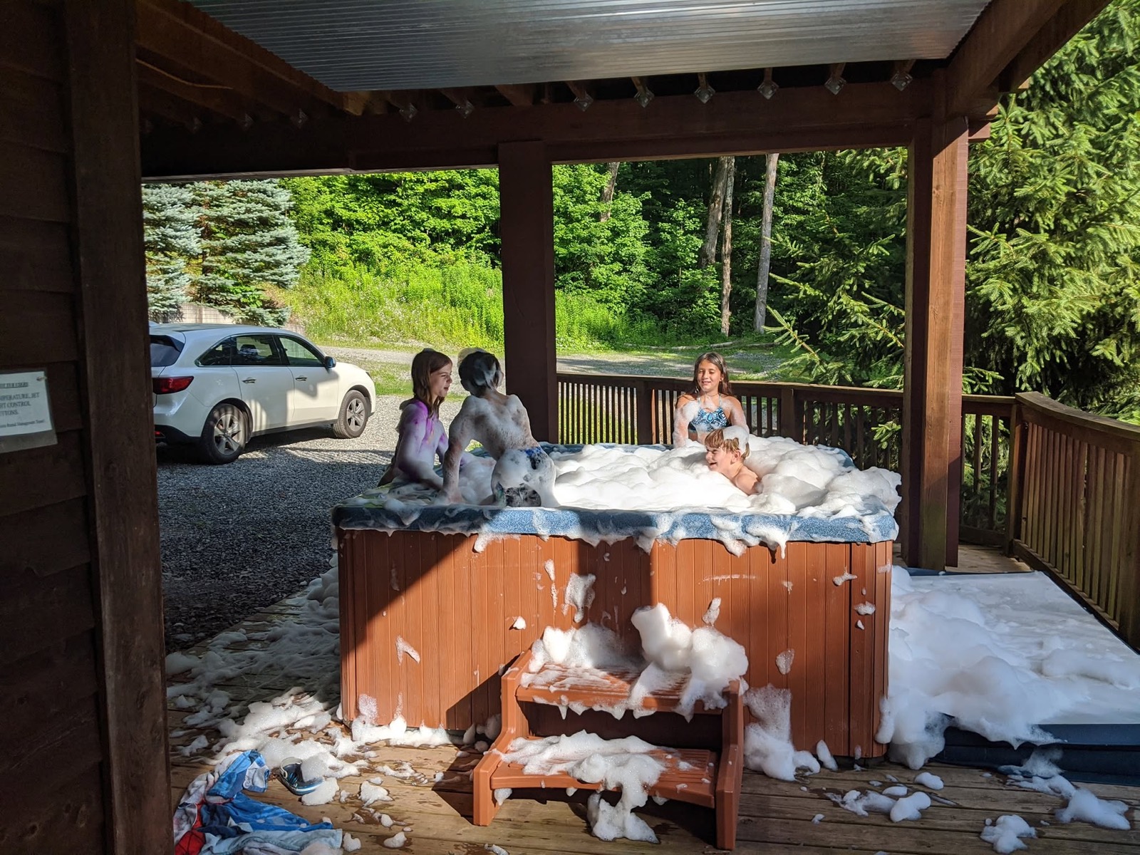 A hot tub on a covered cabin deck erupting with a huge mound of white foam that has spilled out over the sides and across the decking, children watching from the edge.