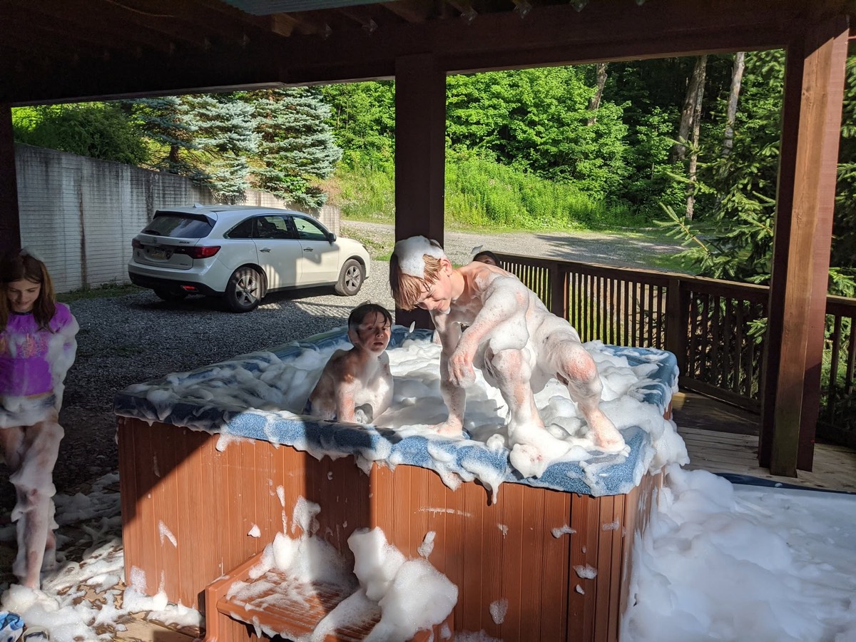 The foam-covered hot tub again with a child climbing up beside it and another standing at the left, suds spread across the deck boards and a car parked beyond the railing.