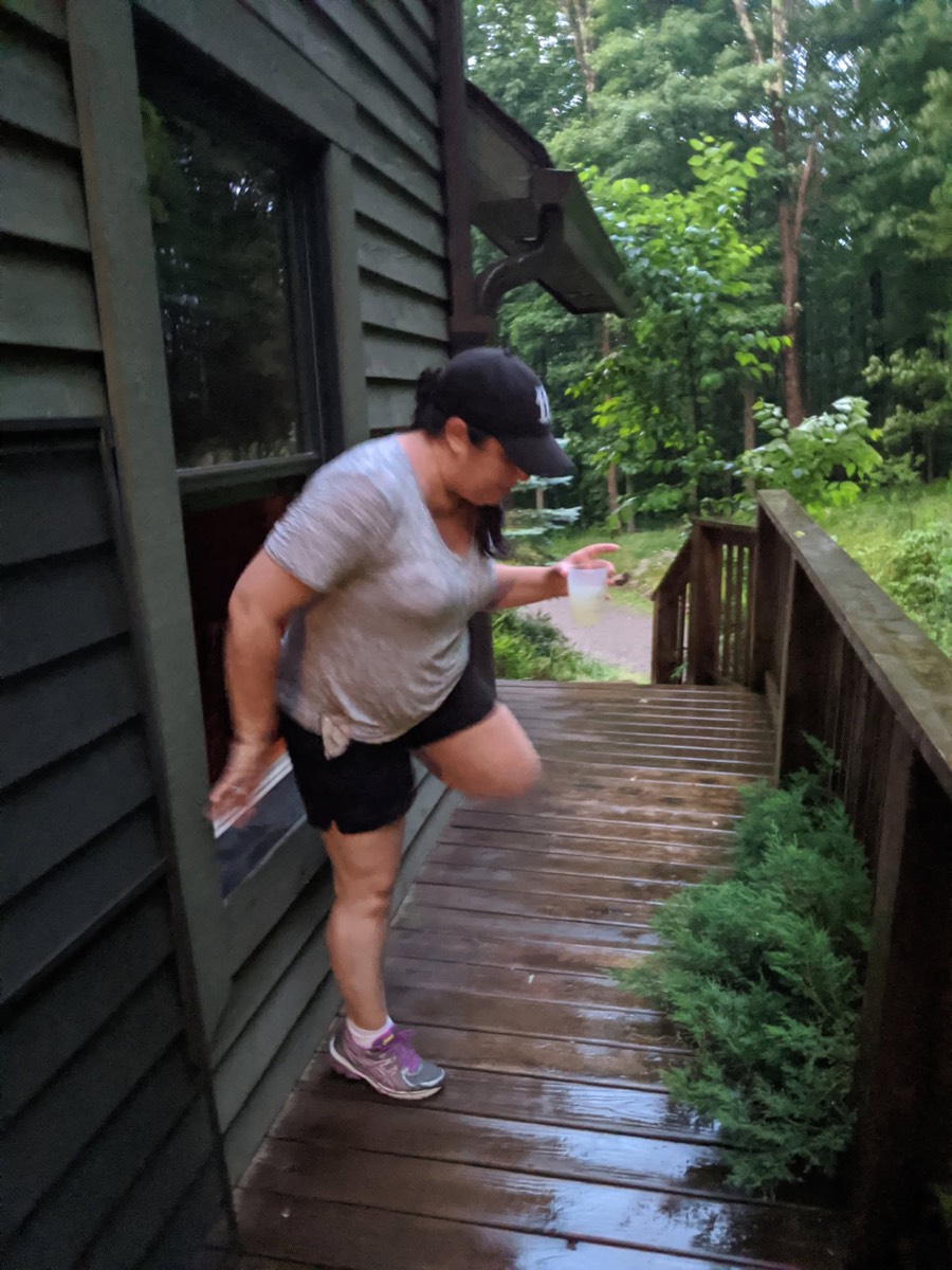 Lanay Zarallo stepping out through a low opening in the cabin wall onto the rain-soaked deck, a drink in one hand and her back leg still raised, wet boards and green woods beyond.