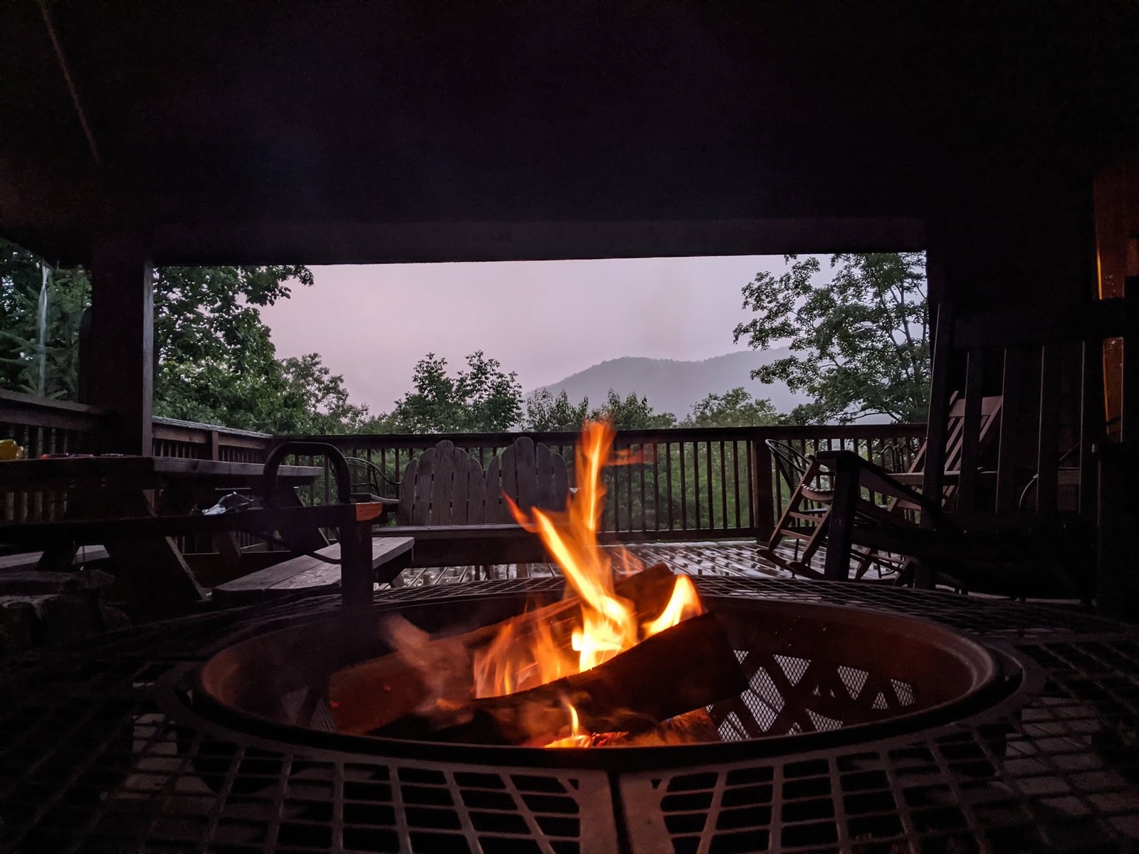 A fire burning in a round deck fire pit at dusk under the cabin's covered porch, chairs pulled around it and a dark blue mountain ridge on the horizon.