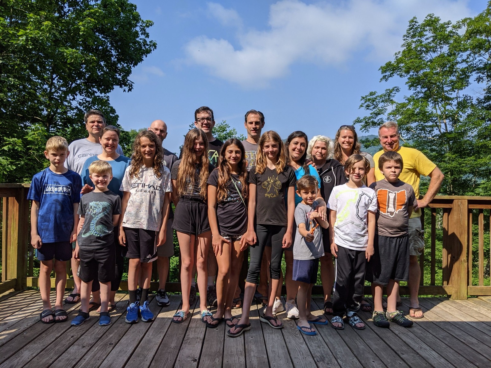 The full Chuckwagon Gang posed on a wooden cabin deck, adults standing in a back row behind nine children, green mountains and a bright blue sky behind them.