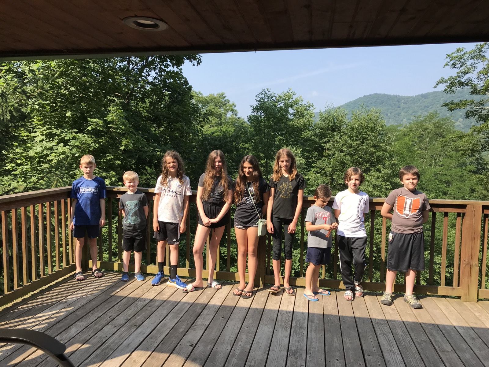 Nine children lined up along a wooden deck railing under a cabin overhang, forested green mountains behind. Left to right: Colton in a blue Unspeakable tee, Owen in a dragon tee, Mason in a white Etihad soccer shirt, Claire in tie-dye, Vivian with a green shoulder bag, Brook in a black and gold tee, Sam in a Phillies shirt, Ryan in white and Asher in an Ohio tee.