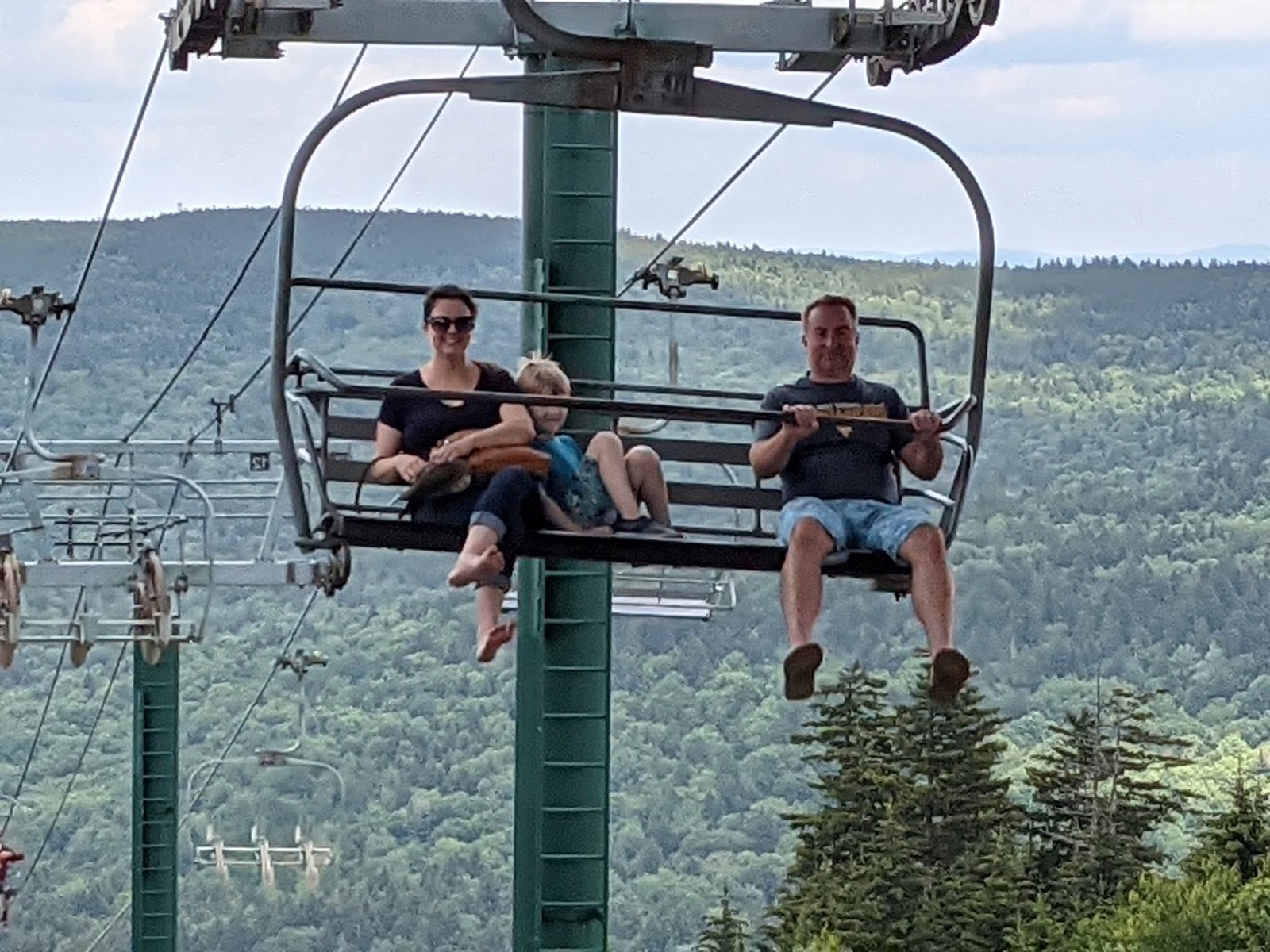 Two adults and a small child riding a green chairlift high above a forested valley, with ridges receding into haze behind them.