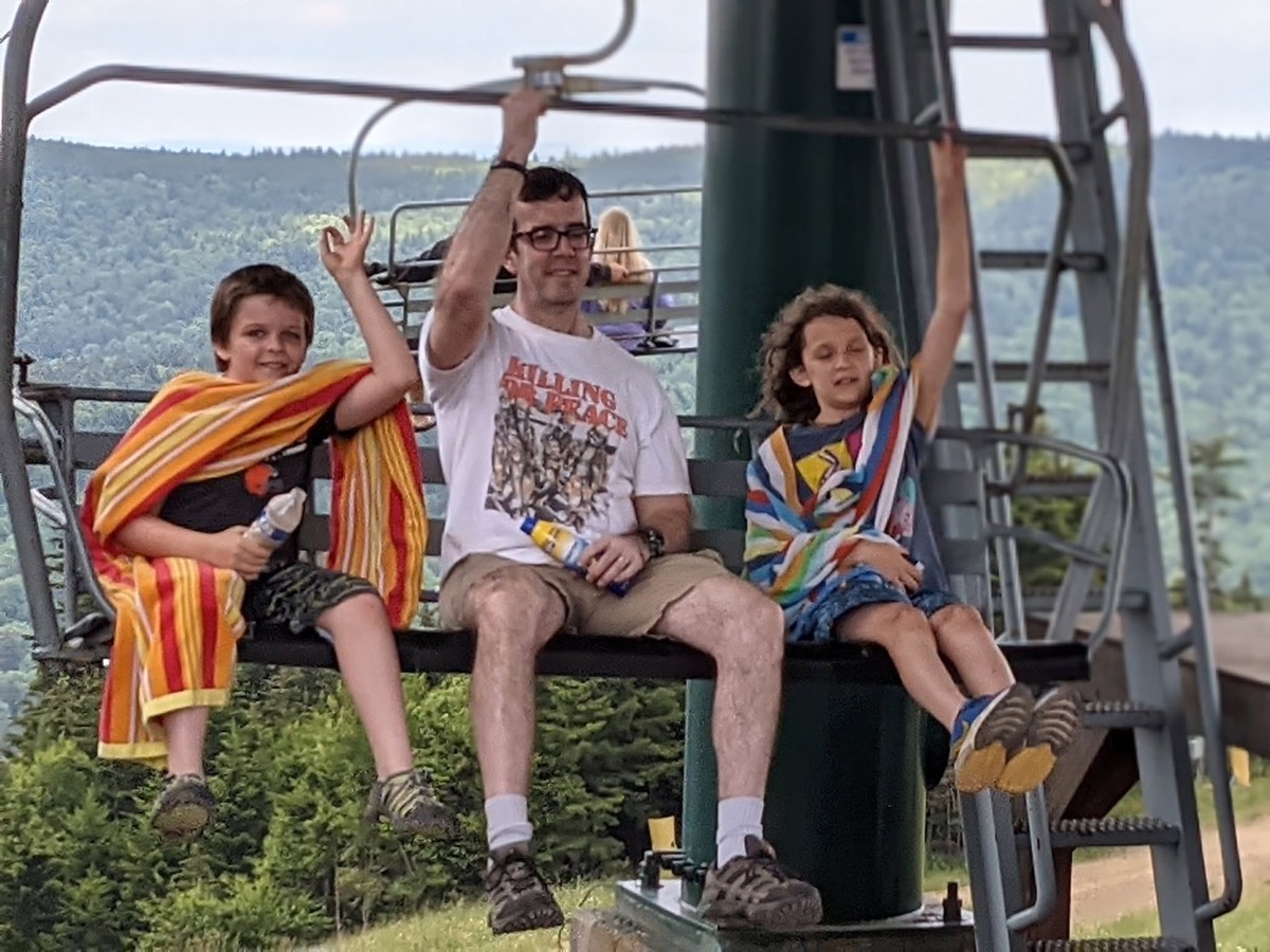 A man and two children riding a chairlift together, one boy in a striped towel raising his arm, wooded mountains behind.