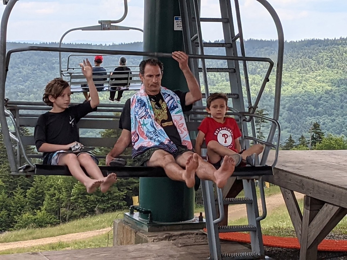 A man and two children on a chairlift approaching the wooden top station platform, green hills beyond.