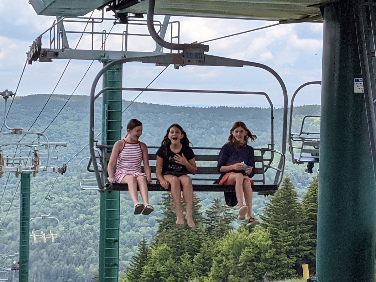 Three girls riding a chairlift together over dark spruce woods, laughing, a line of empty chairs stretching away behind them.