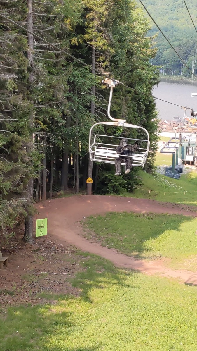 A distant chairlift chair descending over a grassy ski slope and mountain bike trail with a single dark shaggy figure seated in it.