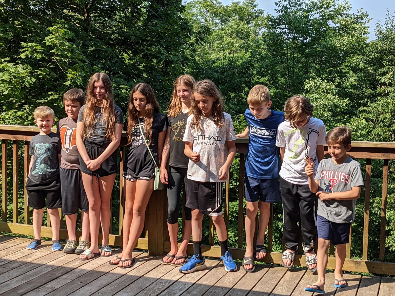 The nine Chuckwagon Gang children shoulder to shoulder along the deck railing in bright sun. Left to right: Owen in a dragon tee, Asher in an Ohio tee, Claire in tie-dye, Vivian with a green shoulder bag, Brook in a black and gold tee, Mason in a white Etihad soccer shirt, Colton in a blue Unspeakable tee, Ryan in white and Sam in a Phillies shirt.