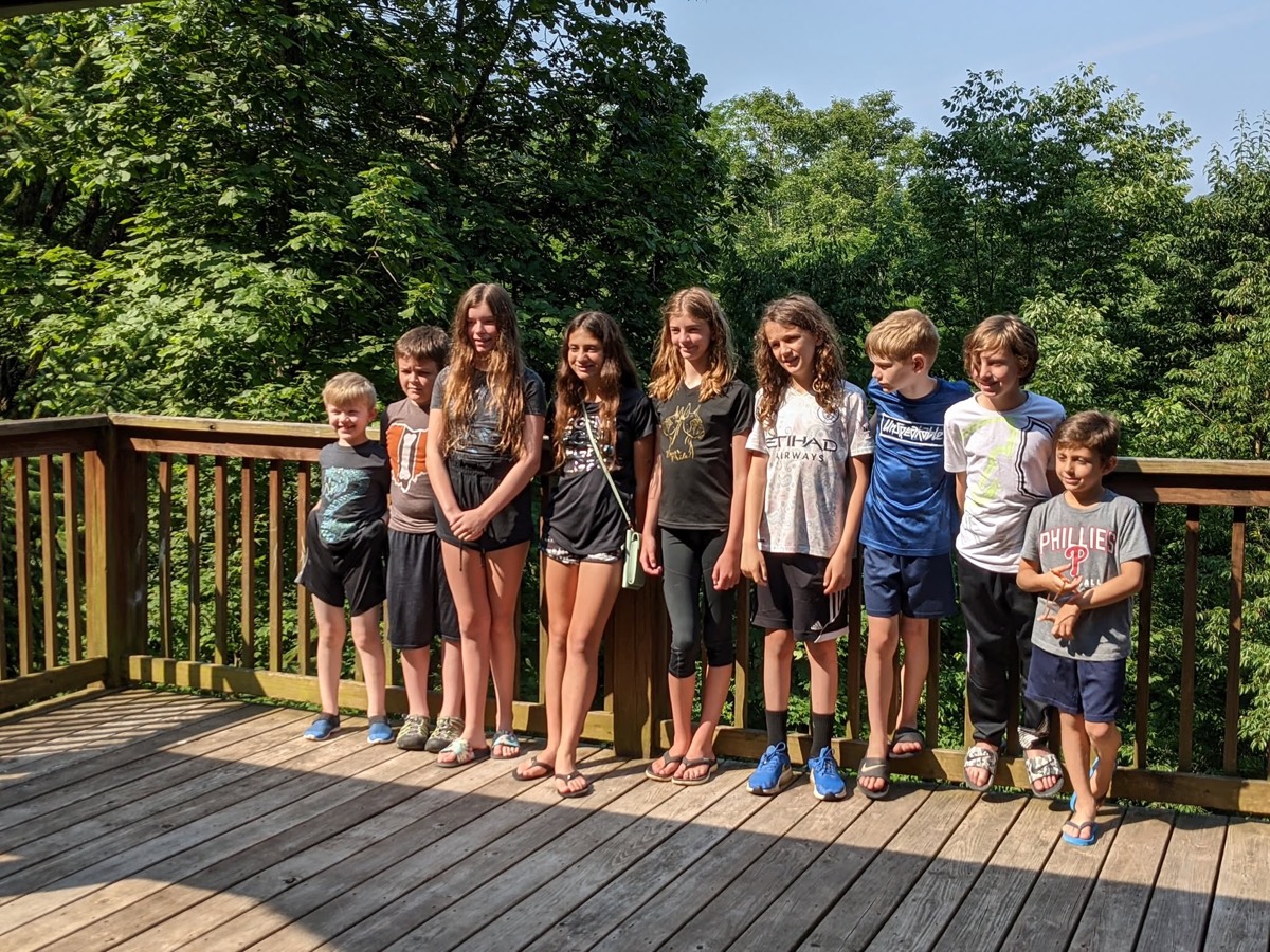 The same row of nine children along the deck railing from further back, the full length of the sunlit deck visible: Owen, Asher, Claire, Vivian, Brook, Mason, Colton, Ryan and Sam.