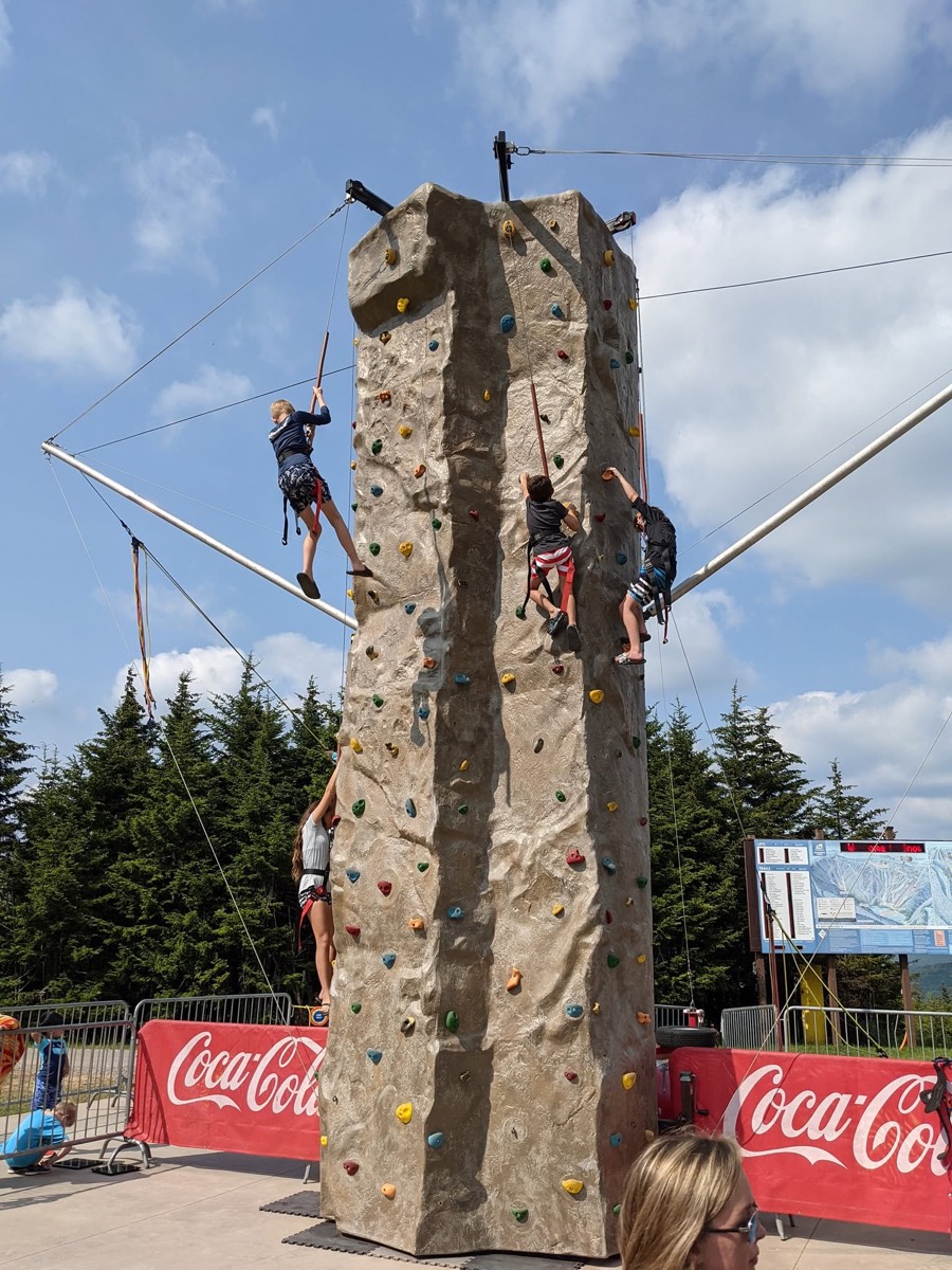 A tall artificial climbing tower with three children partway up different faces on harnesses, Coca-Cola barriers at its base and blue sky behind.