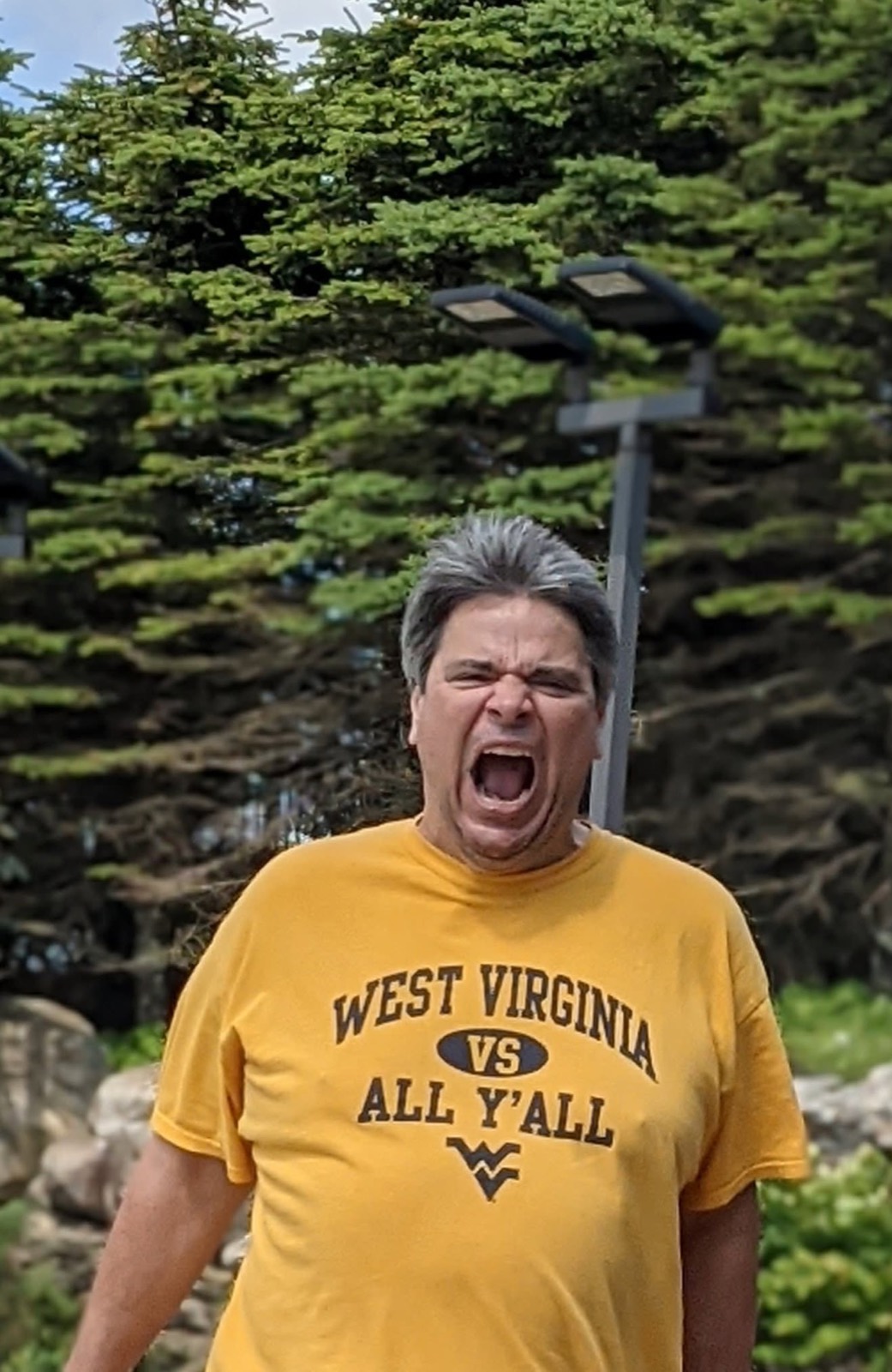George McElwee in a gold West Virginia vs All Y'all t-shirt standing in front of dark spruce trees, mouth wide open mid-yell, eyes shut.